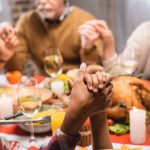 multicultural family holds hands around table and prays before holiday dinner