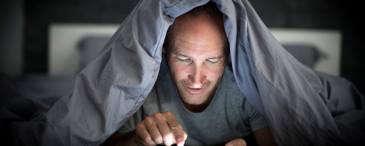 A young cell phone addict man awake late at night in bed using smartphone